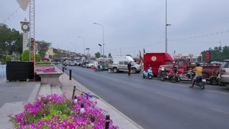 Static-shot-of-a-road-with-vehicles-passing-while-people-wait-to-cross-to-the-other-side-of-the-road-to-get-to-their-parked-vehicles