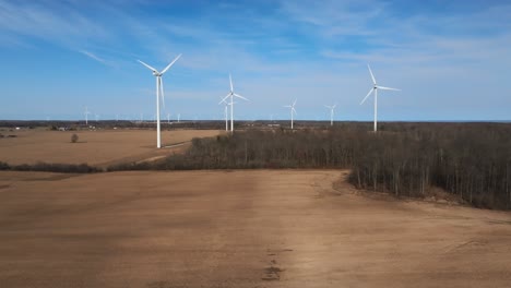 Wind-turbines-spin-in-a-sunny-field,-framed-by-blue-skies-and-a-line-of-trees,-capturing-a-serene-energy-scene