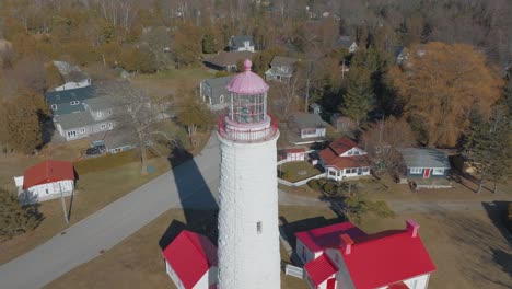 A-charming-lighthouse-with-a-pink-top-surrounded-by-quaint-houses-and-leafless-trees-in-a-coastal-village,-aerial-view