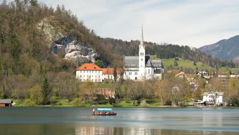 Traditional-Pletna-boat-with-tourists-and-beautiful-church-at-Lake-Bled,-Slovenia