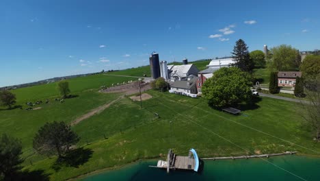 Aerial-approaching-shot-of-pond-at-farmstead-with-silo-storage-and-barn-in-USA