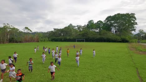 Grupo-De-Escolares-Riendo-Corriendo-Hacia-La-Cámara-En-Un-Campo-De-Fútbol-Verde-Jugando-En-Uniforme-En-La-Selva-Y-El-Bosque-De-Ecuador---Disparo-Revelador-De-Drones