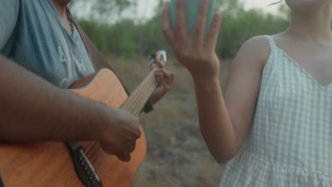 Close-up-of-a-man-playing-guitar-beside-a-woman-in-a-sundress,-outdoors-at-dusk