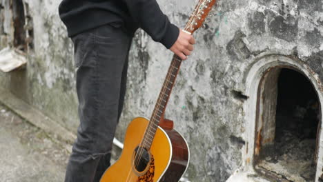 Young-man-wearing-a-black-hoodie-walking-towards-a-classical-guitar,-then-taking-it-with-a-hand-and-holding-it-still,-in-an-abandoned-structure,-on-a-cloudy-day