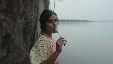 Young-woman-in-traditional-dress-enjoys-a-popsicle-by-the-lake,-with-soft-focus-and-moody-ambiance