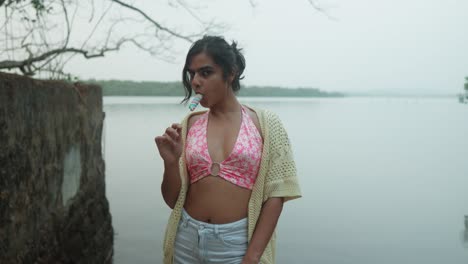 Young-woman-in-summer-attire-enjoys-a-popsicle-by-a-tranquil-lake,-cloudy-day,-relaxed-mood