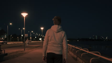 Siga-Hacia-Atrás-Y-Verá-Una-Foto-Panorámica-De-Un-Joven-Que-Comienza-A-Caminar-De-Noche-En-Una-Ciudad-Moderna,-Un-Paisaje-Urbano-Con-La-Carretera-Iluminada-Y-Una-Calle-Bien-Iluminada-Frente-Al-Mar.