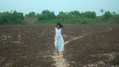 Woman-in-white-dress-walking-on-dirt-path-through-barren-field,-conveying-a-sense-of-solitude-and-reflection