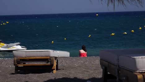 Frau-In-Rot-Sitzt-Am-Lavastrand-In-Perissa,-Santorini,-Griechenland-Und-Genießt-Die-Wärme-Und-Das-Wunderschöne-Meer-An-Der-Küste