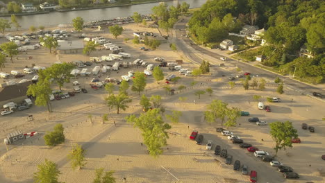 Vista-Aérea-Y-Toma-Panorámica-De-Un-Campamento-Y-Estacionamiento-Con-Autos-Conduciendo-Y-Buscando-Un-Lugar-De-Estacionamiento-Ubicado-Junto-A-Una-Playa-Y-Un-Río