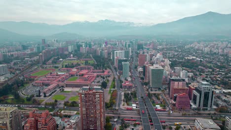 Aerial-Drone-fly-misty-neighborhood-of-Santiago-de-Chile-cordillera-mountain-background,-urban-green-area-around-Escuela-Militar