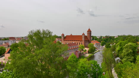 An-aerial-view-of-Lidzbark-Warmiński-showing-the-historic-castle-with-red-tiled-roofs,-surrounded-by-lush-green-trees-and-a-river