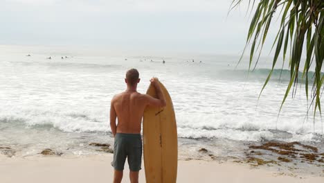 Fittes-Männliches-Model-Steht-Am-Tropischen-Strand-Mit-Surfbrett-Und-Blickt-Auf-Die-Wellen