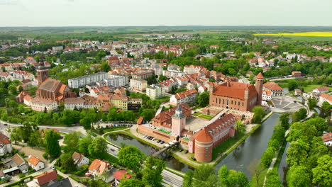 Una-Amplia-Toma-Aérea-De-Lidzbark-Warmiński,-Que-Captura-El-Castillo-Histórico,-El-Río,-Los-Edificios-Circundantes,-Los-árboles-Verdes-Y-Una-Amplia-Vista-De-La-Ciudad-Y-El-Campo.