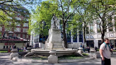 Spring-day-at-Leicester-Square-with-William-Shakespeare-statue-surrounded-by-lush-trees-and-bustling-city-life,-wide-shot