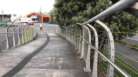 Un-Ciclista-Que-Lleva-Un-Chaleco-De-Alta-Visibilidad-Recorre-Un-Puente-Peatonal-Hacia-Una-Calle-Muy-Transitada-En-Auckland,-Nueva-Zelanda