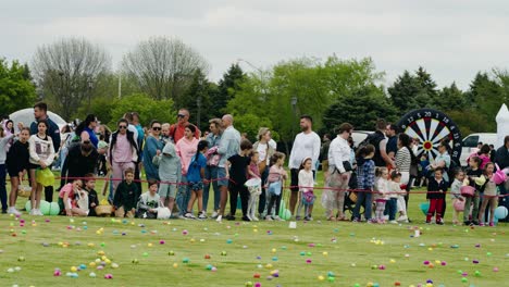 Parents-and-children-lined-up-on-a-grassy-field,-ready-for-an-egg-hunting-event-filled-with-colorful-eggs