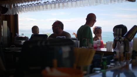 Bar-by-the-beach-on-a-bright-sunny-day-with-people-having-drinks-and-enjoying-the-sun-on-the-European-Coast,-handheld-shot