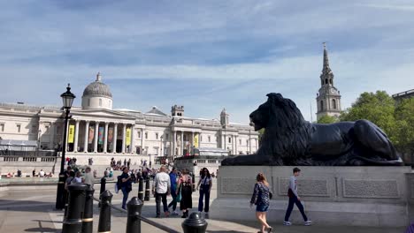 Sunny-day-at-Trafalgar-Square-with-tourists-near-the-iconic-lion-statue,-National-Gallery-in-background