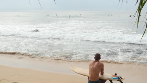 Man-Sitting-on-Sandy-Beach-With-Surfboard-Looking-Out-Over-the-Waves