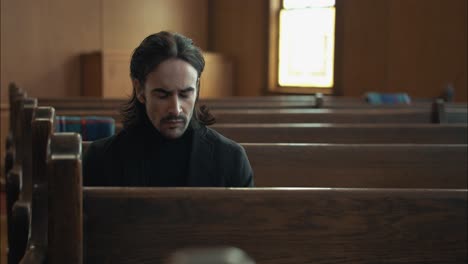 Young-man-with-dark-hair,-emotional,-sombre-man-in-black-suit-sitting-in-pew-in-church-in-cinematic-slow-motion