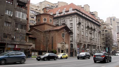 City-traffic-flows-past-old-and-modern-buildings-on-a-cloudy-day-in-Bucharest,-Romania,-Biserica-Italiana