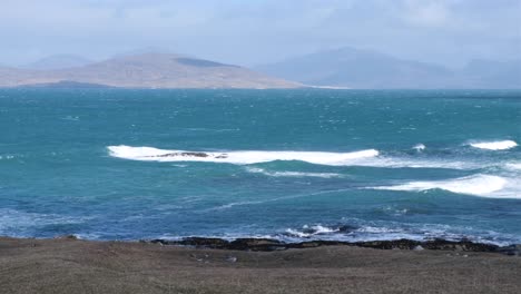 Scenic-view-of-wild-waves-rolling-in-on-turquoise-blue-ocean-and-mountains-in-the-distance-on-isle-of-Lewis-and-Harris-in-Outer-Hebrides,-Western-Scotland-UK