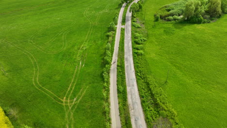 Aerial-view-of-a-rural-road-with-two-bikers-riding-along-it,-bordered-by-lush-green-fields-with-visible-tire-tracks