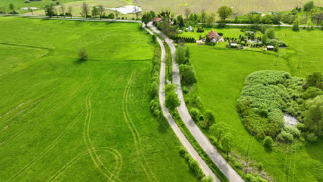 Aerial-view-of-a-winding-rural-road-through-green-fields-and-small-settlements,-with-farmlands-in-the-background