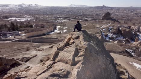 Cappadocia-Adventure:-Solo-Tourist-Hiker-Sits-Atop-Mountain-Cave-Dwelling-Enjoying-View-Towards-Göreme-and-Uchisar-Castle-on-Horizon