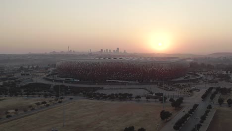 Luftaufnahme-Einer-Drohne-Im-Orbit-Des-FNB-Stadions-Bei-Sonnenaufgang-Mit-Johannesburg-CBD-In-Der-Ferne_01