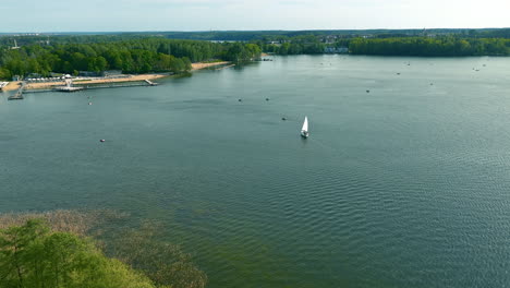 A-wide-view-of-Ukiel-Lake,-featuring-a-single-sailboat-and-a-sandy-beach-area-on-the-left