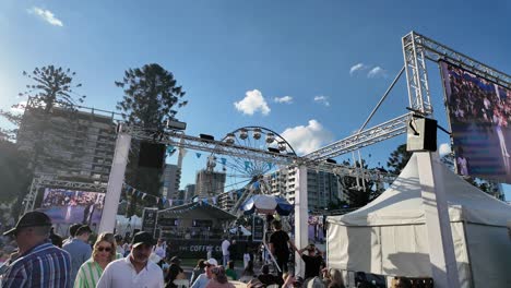 Gente-Relajándose-En-El-Festival-Griego-Paniyiri-Con-Escenario-Y-Noria-Al-Fondo