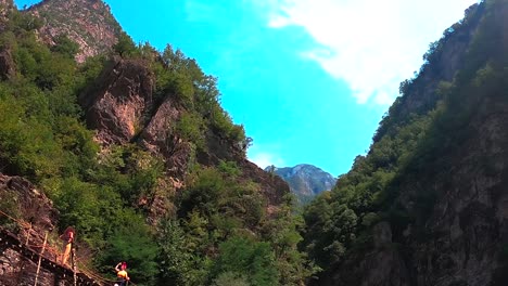 Paradise-Alpine-landscape-with-broken-wooden-bridge-over-crystal-water-of-river-surrounded-by-rocky-mountains-under-bright-sky