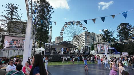 Niños-Jugando-En-El-Festival-Griego-Paniyiri-Con-Escenario-Y-Noria-Al-Fondo.