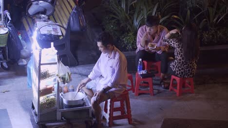 Vendedor-De-Comida-Callejera-Asiática-Cocinando-Con-Una-Pareja-Comiendo-En-Sillas-De-Plástico-Rojo-Por-La-Noche