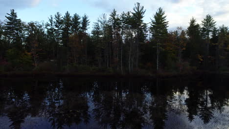 Toma-De-Camión-De-Izquierda-A-Derecha-De-Una-Costa-Oscura-Y-Cambiante-Llena-De-árboles-Y-Su-Reflejo-En-Un-Lago-Tranquilo-En-Maine-Durante-El-Otoño.