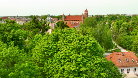 close-up-aerial-view-focusing-on-a-historical-building-with-red-rooftops,-surrounded-by-lush-green-trees-and-other-urban-buildings-in-Lidzbark-Warmiński