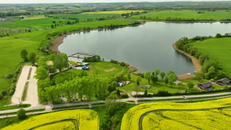 An-aerial-view-of-Wielochowskie-Lake-surrounded-by-green-fields-and-yellow-rapeseed-fields,-with-a-road-running-alongside-the-lake