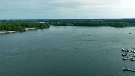 An-aerial-view-of-Ukiel-Lake-in-Olsztyn,-with-boats-and-yachts-on-the-water,-surrounded-by-green-forests-and-a-distant-shoreline