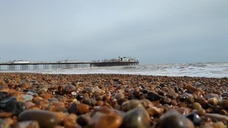 Niedrige-Ansicht-Des-Brighton-Pier-Mit-Kieselsteinen-Im-Vordergrund-Und-Wellen,-Die-In-Zeitlupe-Brechen