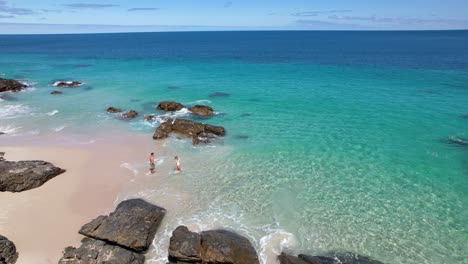 A-couple-holding-hands-and-walking-out-into-bright-blue-crystal-clear-ocean-water-from-a-white-sand-beach