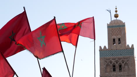 Banderas-Rojas-Marroquíes-Ondeando-Con-La-Mezquita-Koutoubia-En-Marrakech-Al-Fondo,-Luz-Vibrante