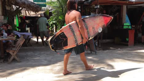 A-tanned-hippie-male-surfer-with-a-red-worn-out-surf-board-walking-barefoot-on-muddy-dirt-road-in-Mexico-after-catching-waves