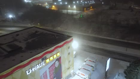 Aerial-drone-shot-descending-down-onto-a-commercial-U-Haul-building-during-a-snowstorm,-rental-vehicles-at-a-standstill-due-to-the-treacherous-weather-conditions