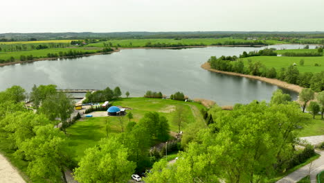 Una-Vista-Aérea-Del-Lago-Wielochowskie-Rodeado-De-Campos-Verdes,-árboles-Y-Una-Pequeña-Zona-Recreativa-Con-Algunos-Edificios