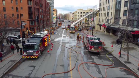 Aerial-view-of-firefighters-and-fire-trucks-on-the-street-extinguishing-a-fire-on-a-building,-flyover