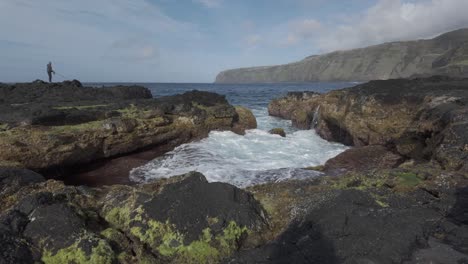 Rugged-coastline-of-Mosteiros,-São-Miguel,-with-waves-crashing-over-black-volcanic-rocks,-hiker-in-distance