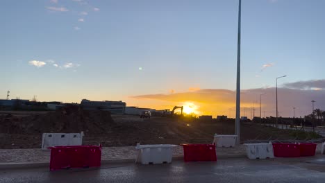 Construction-site-and-car-passing-at-sunset,-escavator