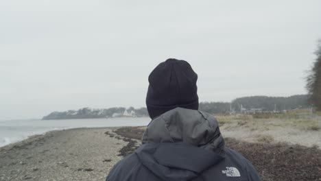 Guy-walks-on-beach-next-to-water-on-windy-and-cloudy-day,-tracking-shot